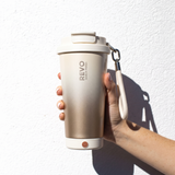 A hand holding a metallic beige REVO glass tumbler with a white lid and wrist strap against a white textured wall.