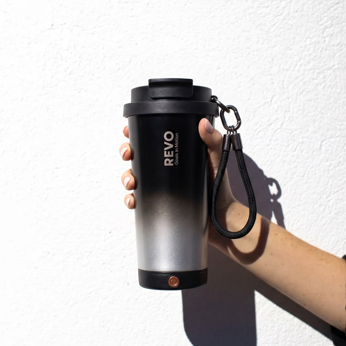 A hand holding a black and silver gradient insulated travel tumbler with a black lid, carrying strap, and visible REVO branding, set against a white wall with a strong shadow.