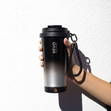 A hand holding a black and silver gradient insulated travel tumbler with a black lid, carrying strap, and visible REVO branding, set against a white wall with a strong shadow.