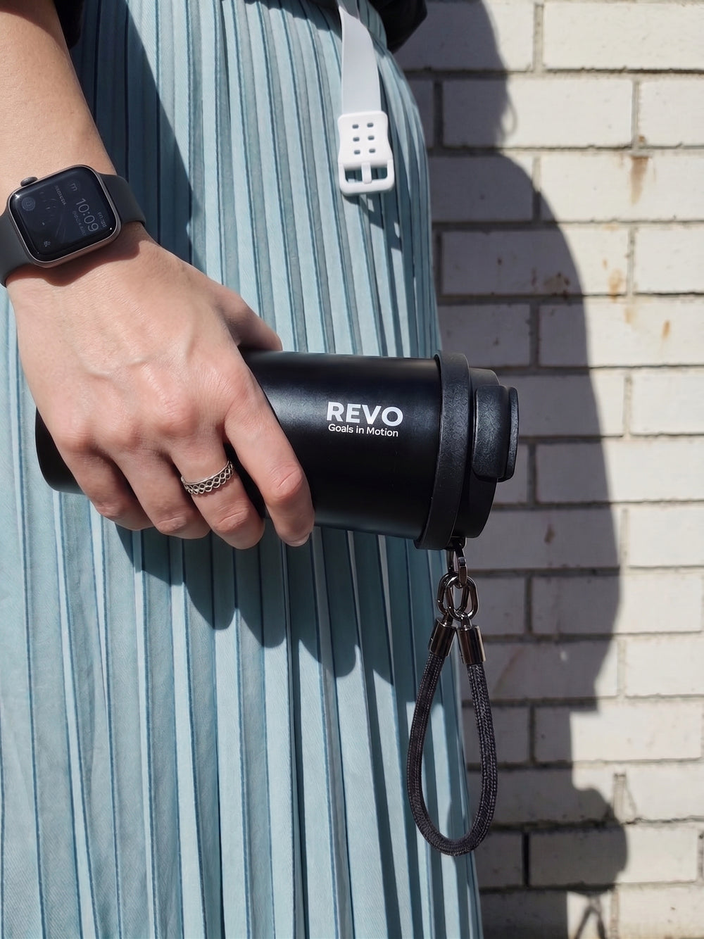 A person in a pleated light blue dress holds a black REVO glass to-go mug with a matching black strap against a white brick wall.