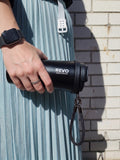 A person in a pleated light blue dress holds a black REVO glass to-go mug with a matching black strap against a white brick wall.